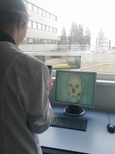 Sieviete stāv ar muguru un skatās uz monitoru, kurā redzams galvaskauss, aiz monitora ir logs ar skatu uz ēku