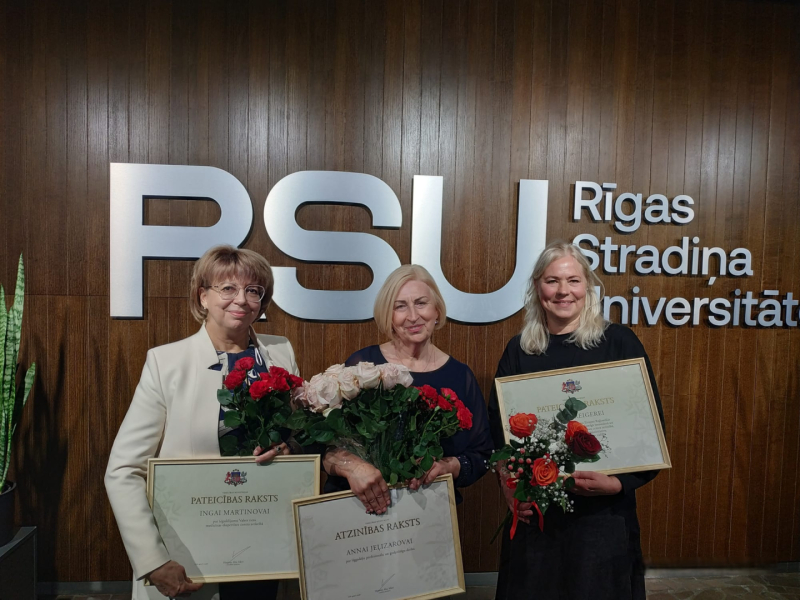 Ingа Martinova, Anna Jeļizarova un Ilze Kreigere ar Veselības ministrijas apbalvojumiem un ziediem pie RSU sienas logo