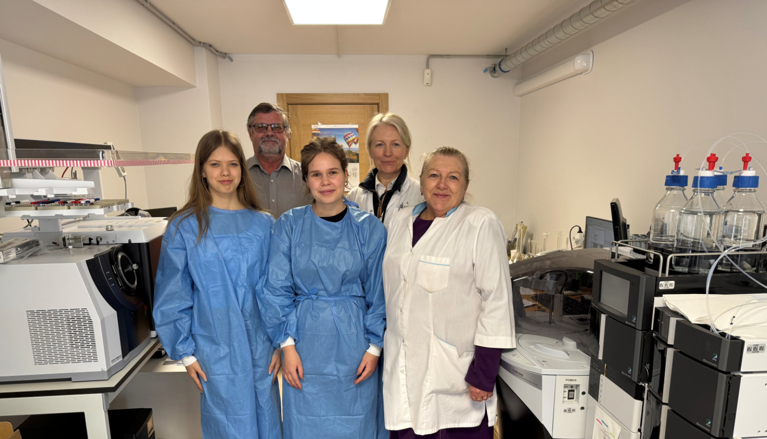 Baltā telpā ar laboratorijas iekārtām stāv pieci cilvēki, divas sieviete baltos halātos, viens vīrietis ar brillēm un divas jaunietes zilos halātos