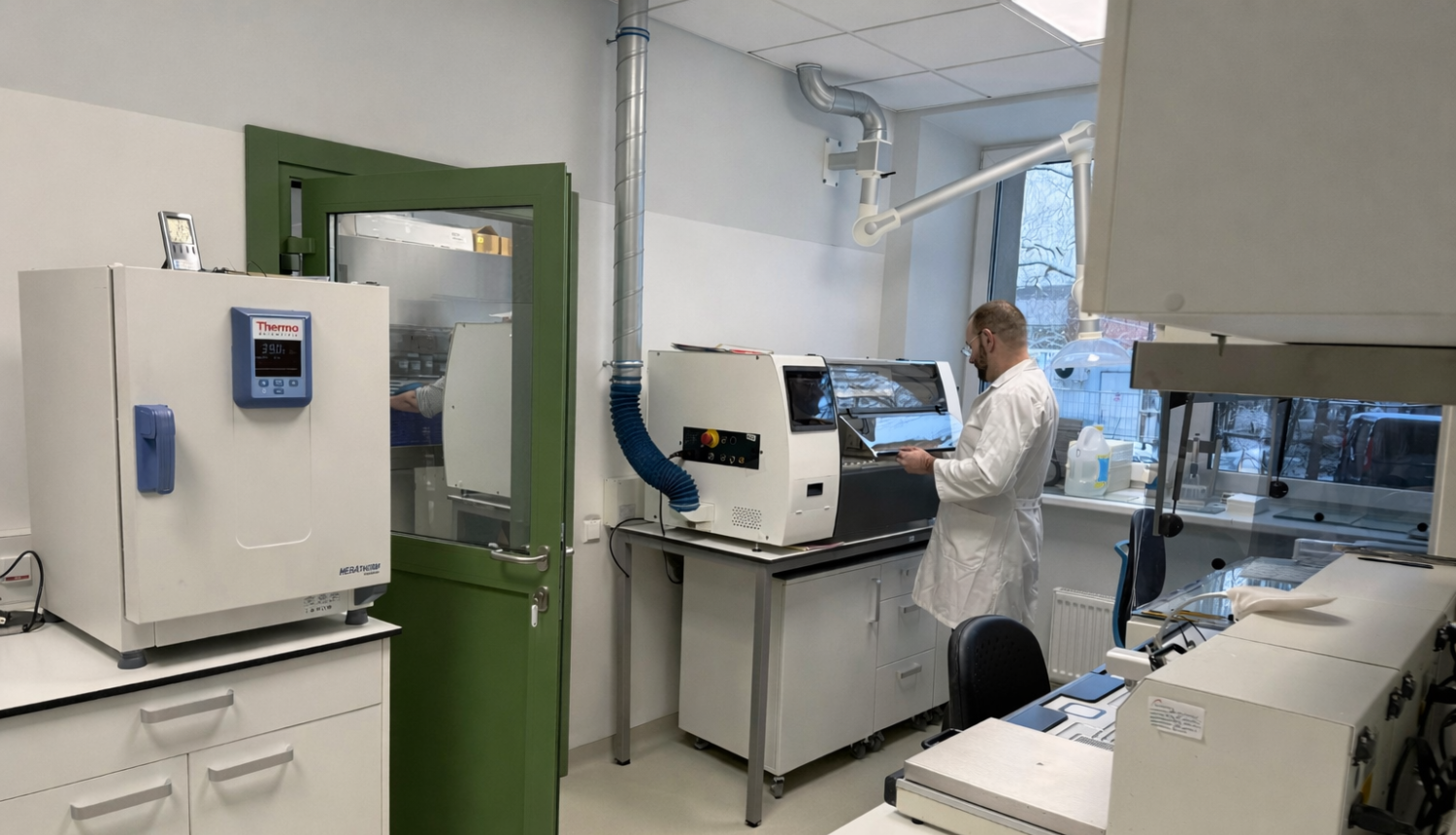 Telpa ar baltām sienām un zaļām durvīm, telpā atrodas laboratorijas iekārtas, vīrietis baltā halātā stāv ar muguru pie iekārtām