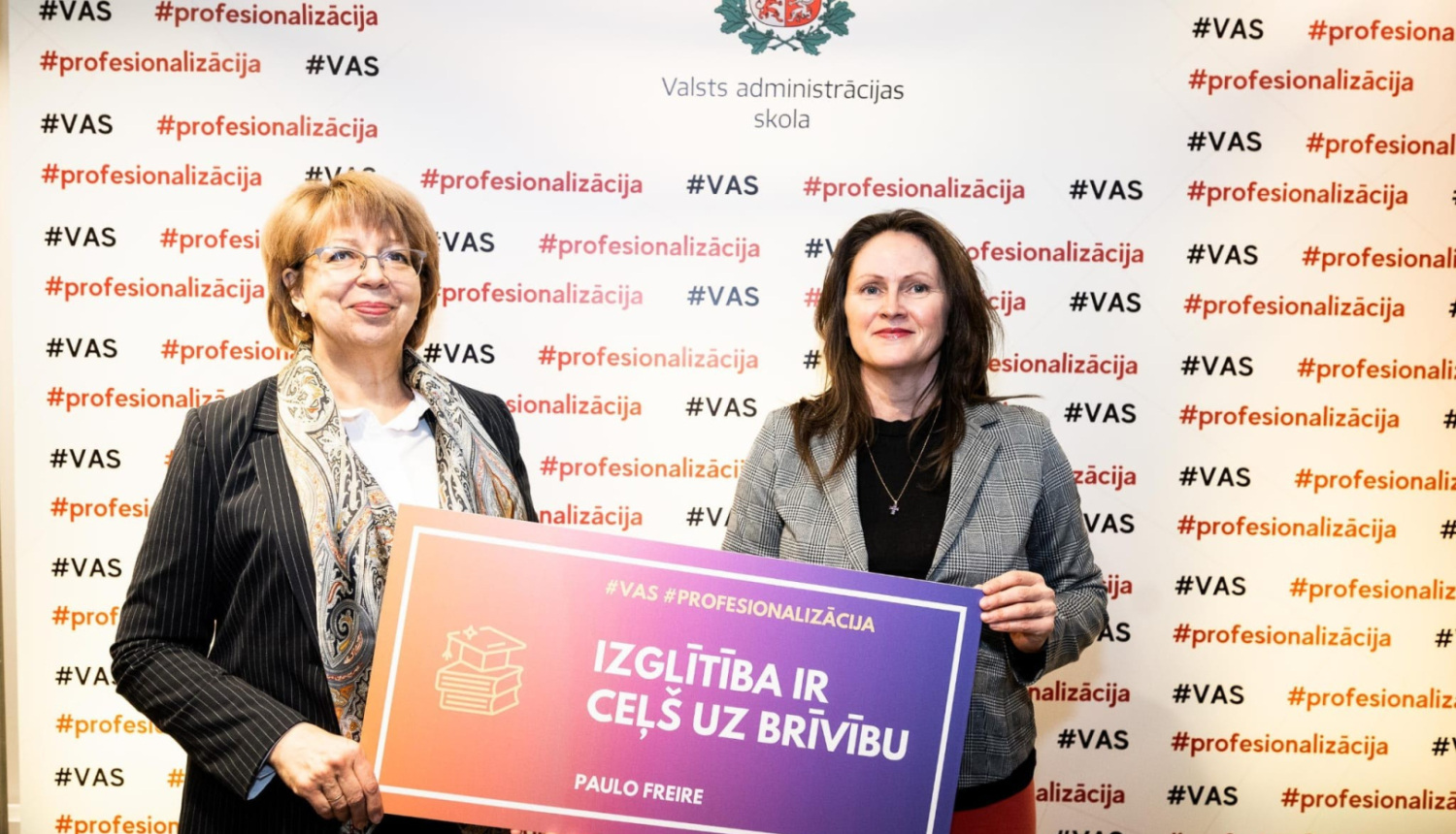 Divas sievietes stāv ar muguru pret sienu, uz kura ģērbonis un uzraksts Valsts administrācijas skola, abas rokā tur plakātu ar uzrakstu - Izglītība ir ceļš uz brīvību