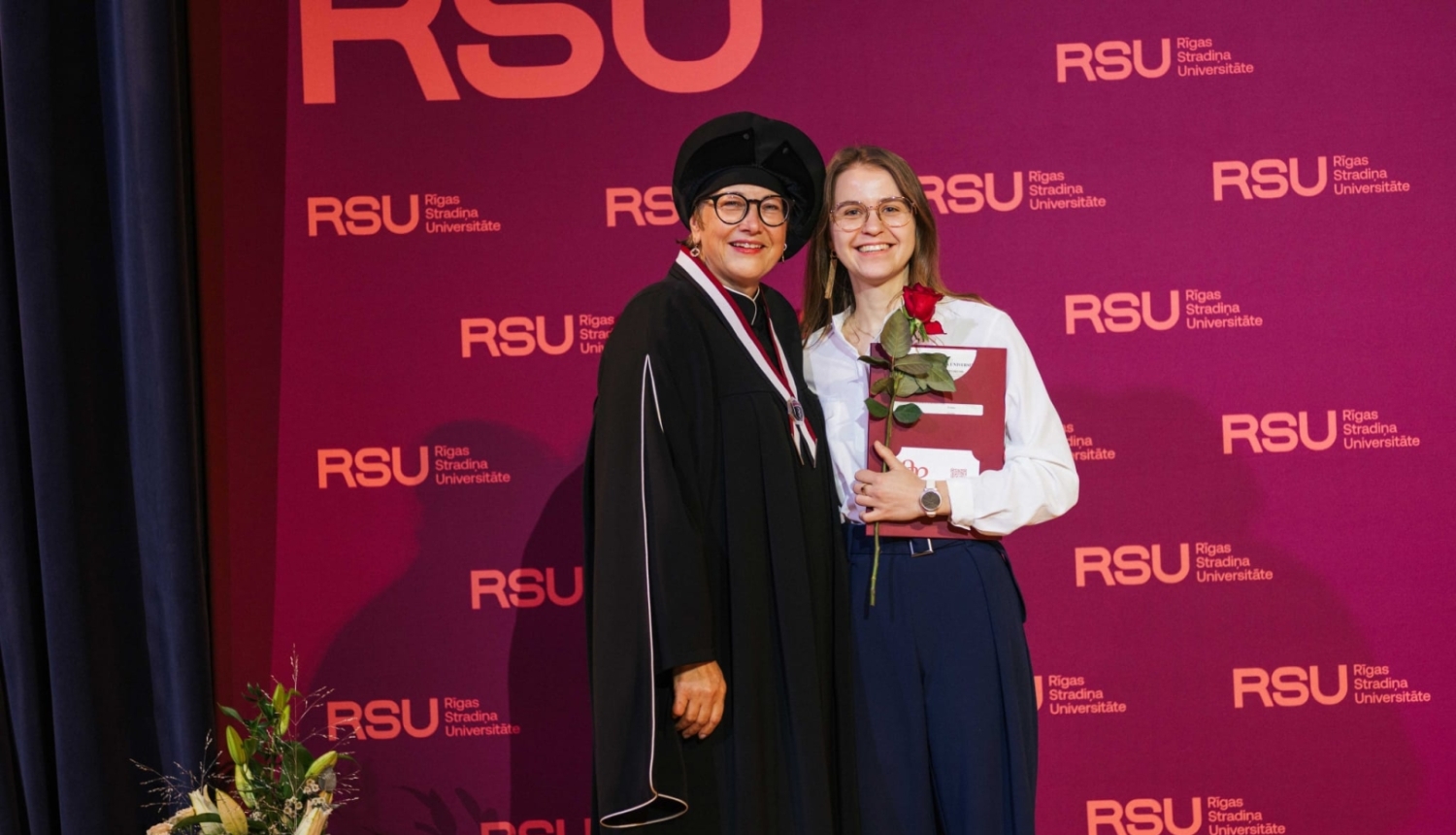 Divas sievietes RSU pasākumā pozē ar diplomu un ziediem.