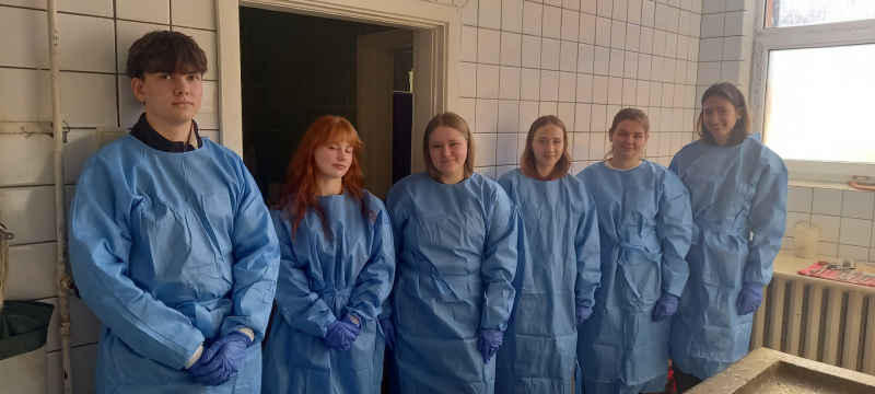 Seši jaunieši zilos halātos stāv pie baltas flīžu sienas