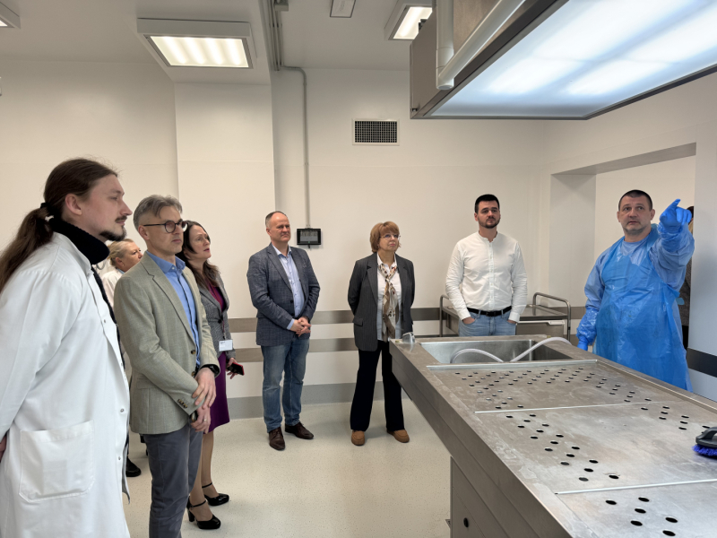 Vairāk cilvēki stāv skciju zālē, apkārt tukšam metāla sekcijas galdam, viens vīrietis zilā halātā kaut ko rāda ar roku