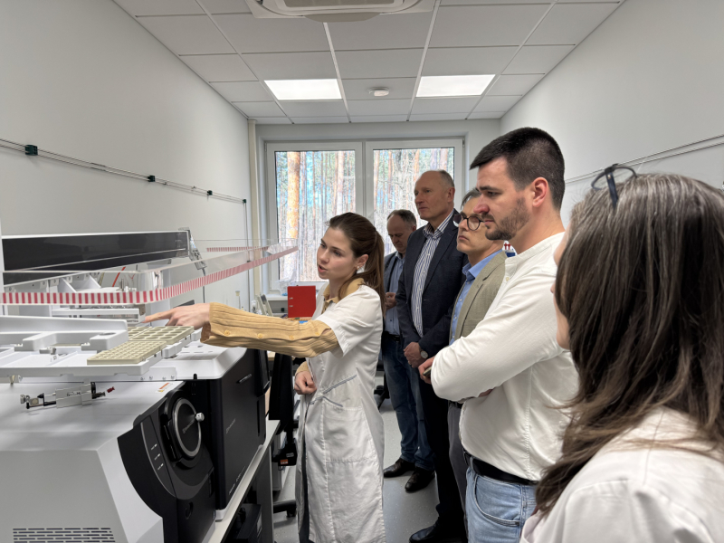 Sieviete baltā halātā darbojas pie laboratorijas iekārtas, vairāki cilvēki aiz viņas stāv un vēro viņas darbības