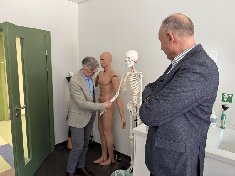 Divi vīrieši stāv kabinetā ar baltām sienām un zaļām durvīm, viens vīrietis tur skeleta roku, balkus tam stāv plastmasas manekens