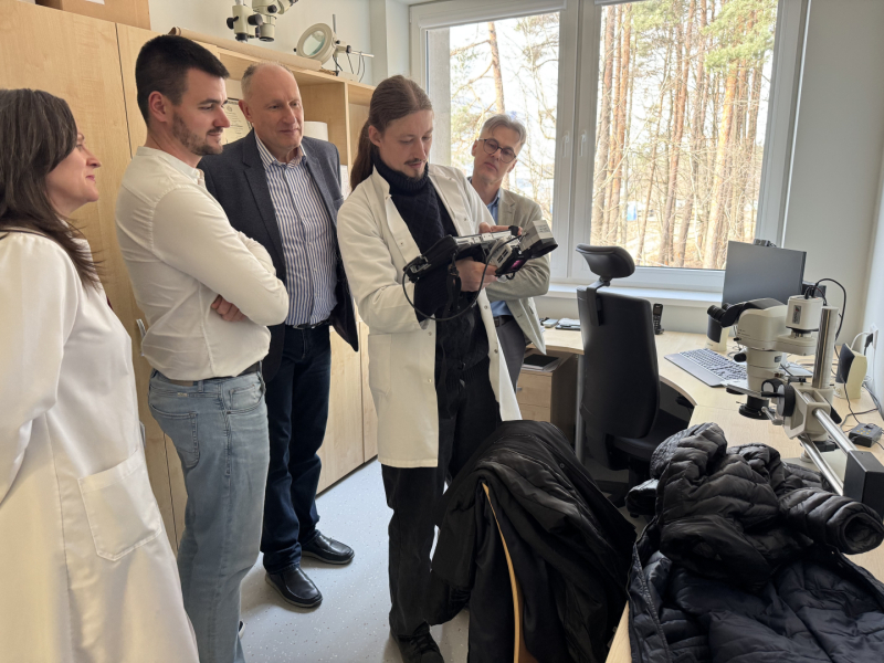 Vairķi cilvēki stāv kabinetā apkārt vīrietim baltā halātā, kas tiem rāda kaut ko laboratoriskā aparātā, kas atrodas viņam rokās