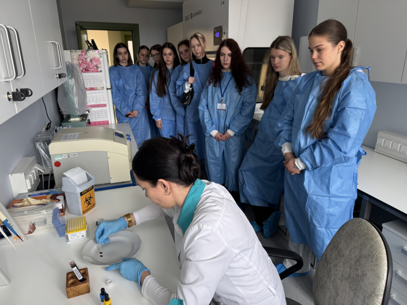 Sieviete baltā halātā sēž pie balta galda uz kura stāv dažādi laboratorijas piederumi, vairāki jaunieši zilos halātos stāv un vēro sievietes darbības