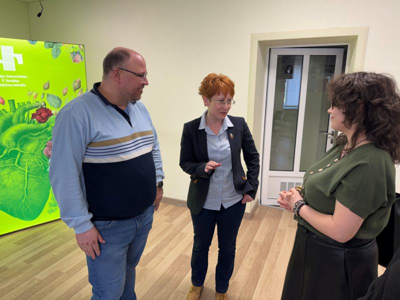 Gaišā auditorijā 2divas sievetes un viens vīrietis stāv un sarunājas