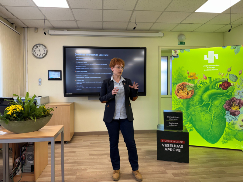 Gaišā auditorijā sieviete stāv un kaut ko stāsta, aiz muguras viņai liels ekrāns ar prezentāciju