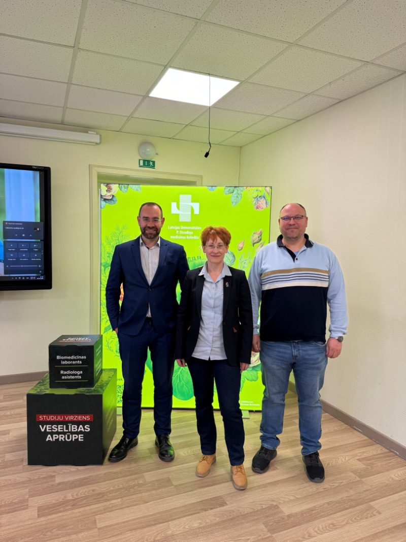 Gaišā auditorijā viena sieviete un divi vīrieši tāv, fonā spilgti zaļš baneris