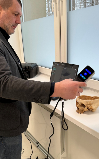 Vīrieti stāv ar portatīvu skeneri rokās pie loga ar baltu palodzi, uz palodzes atrodas apakšējā puse no galvaskausa