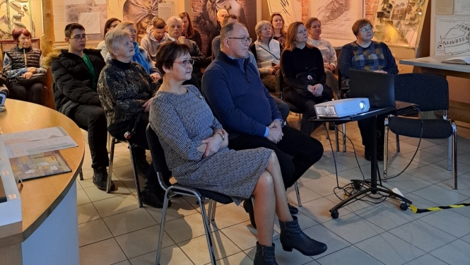 Telpa ar krēsliem rindās un uz tiem sēdošiem cilvēkiem