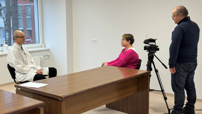 Telpa ar baltām sienām, pie darba galda viens otram pretī sēž vīrietis un sieviete, vēl viens vīrietis stāv un filmē sedošos