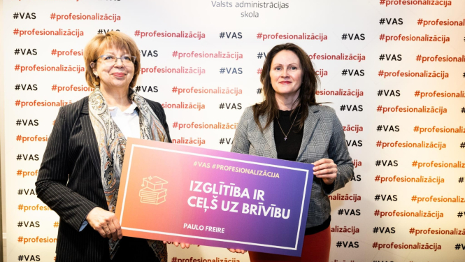 Divas sievietes stāv ar muguru pret sienu, uz kura ģērbonis un uzraksts Valsts administrācijas skola, abas rokā tur plakātu ar uzrakstu - Izglītība ir ceļš uz brīvību