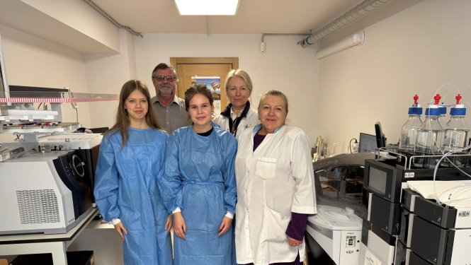 Baltā telpā ar laboratorijas iekārtām stāv pieci cilvēki, divas sieviete baltos halātos, viens vīrietis ar brillēm un divas jaunietes zilos halātos