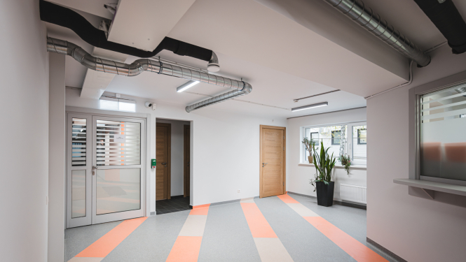 Moderna iestādes iekštelpa ar košu grīdas segumu, koka durvīm un lieliem logiem; redzami gaisa ventilācijas cauruļvadi un zaļie telpaugi pie loga