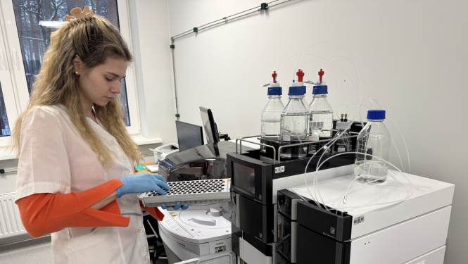 Jauna sieviete baltā halātā un zilos gumijas cimdos stāv laboratorijas telpā, rokās tur laboratorijas piederumus