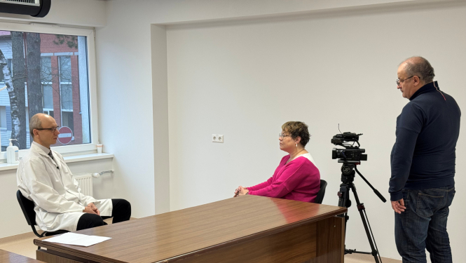 Telpa ar baltām sienām, pie darba galda viens otram pretī sēž vīrietis un sieviete, vēl viens vīrietis stāv un filmē sedošos