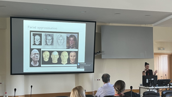 Auditorija ar dalībniekiem pie galdiem un prezentāciju uz ekrāna