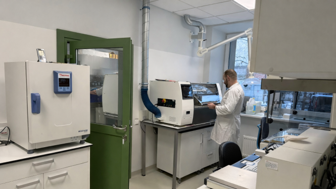 Telpa ar baltām sienām un zaļām durvīm, telpā atrodas laboratorijas iekārtas, vīrietis baltā halātā stāv ar muguru pie iekārtām