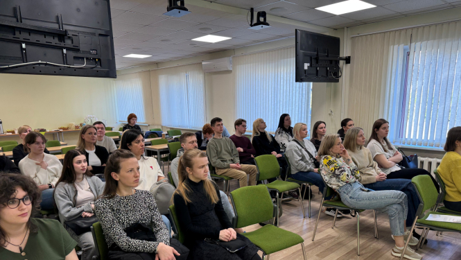 Gaiša auditorija, rindās zaļi krēsli uz kuriem sēž cilvēki