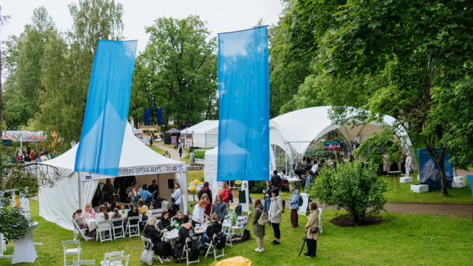 Uz zālāja baltas teltis pie kurām pulcējas cilvēki, v;el uz zālāja balti krēsli, dzelteni sēžammaisi un divi zili karogi