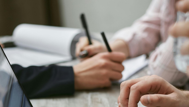 Darba galds ar portatīvo datoru, kā arī redzamas rokas, kas tur pildspalvas