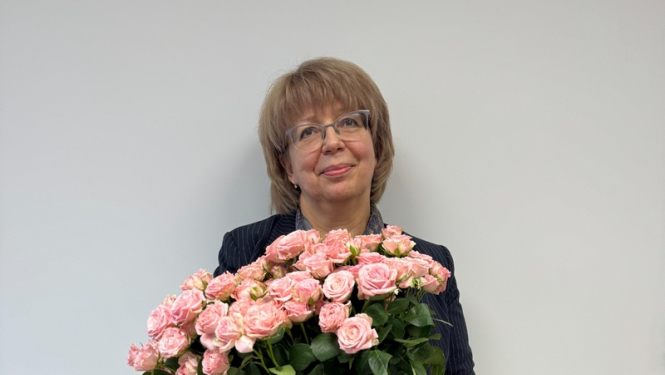 Sieviete stāv un smaida, balta siena fonā, rokās rozā rozes