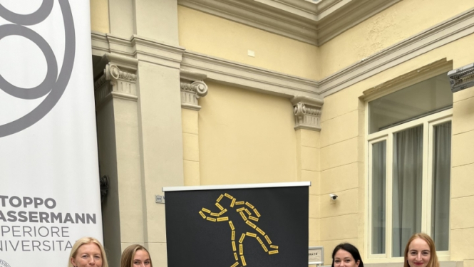 Četras sievietes stāv pie plakāta ar konferences programmu, fonā dzeltena ēka