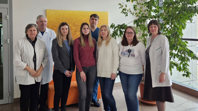 Astoņi cilvēki pozē fotogrāfam, fomā balta siena, liela glezna dzeltenos toņos un liels istabas augs podā