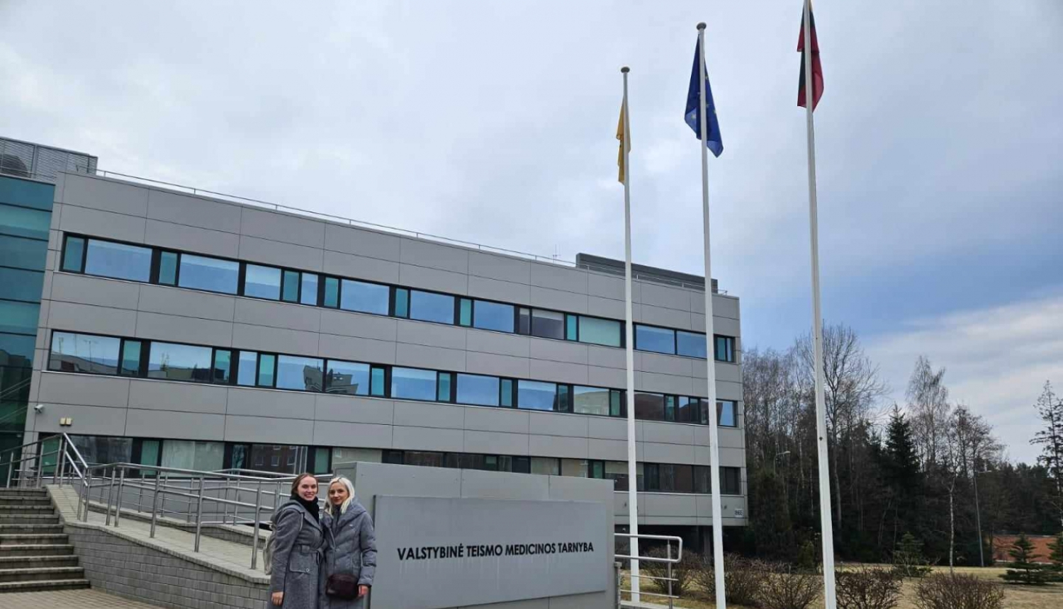 Divas sievietes stāv, fonā pelēka trīs stāvu ēka