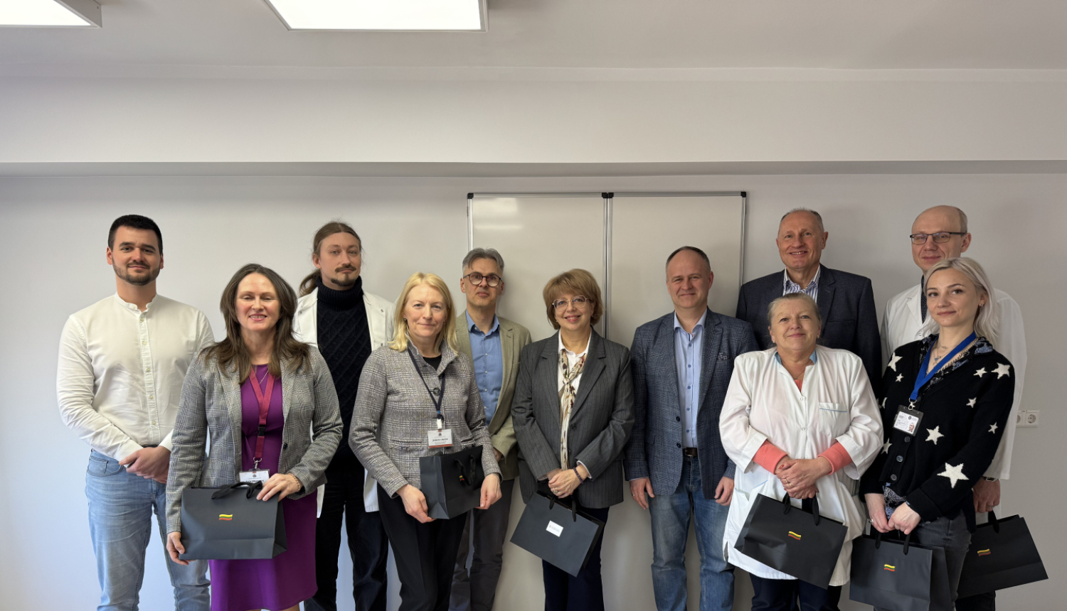 Vienpadsmit cilvēki stāv baltā telpā, vairākiem ir rokās melns dāvanu maisiņš, tiem aizmugurē pie sienas balta tāfele