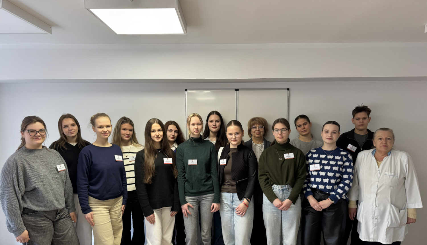 Vairāki cilvēki stāv pie baltas sienas un pozē fotogrāfam