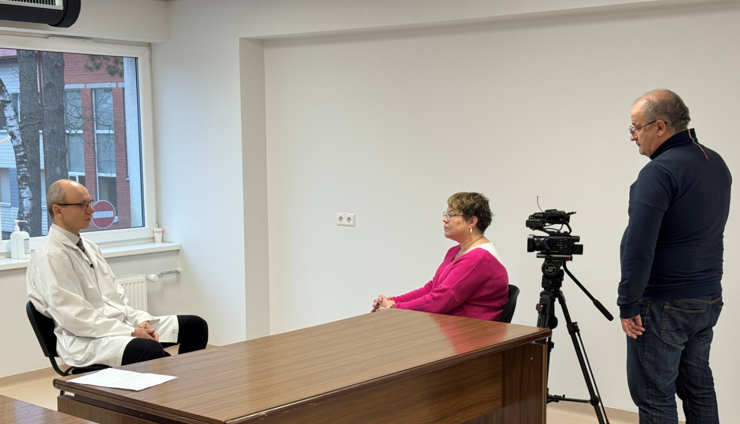 Telpa ar baltām sienām, pie darba galda viens otram pretī sēž vīrietis un sieviete, vēl viens vīrietis stāv un filmē sedošos