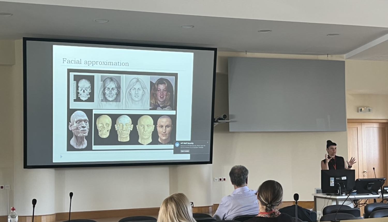 Auditorija ar dalībniekiem pie galdiem un prezentāciju uz ekrāna