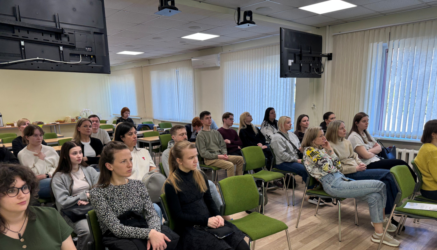 Gaiša auditorija, rindās zaļi krēsli uz kuriem sēž cilvēki