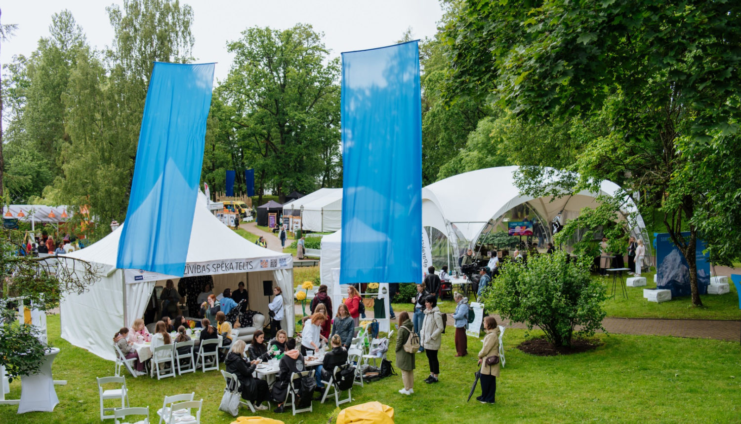Uz zālāja baltas teltis pie kurām pulcējas cilvēki, v;el uz zālāja balti krēsli, dzelteni sēžammaisi un divi zili karogi