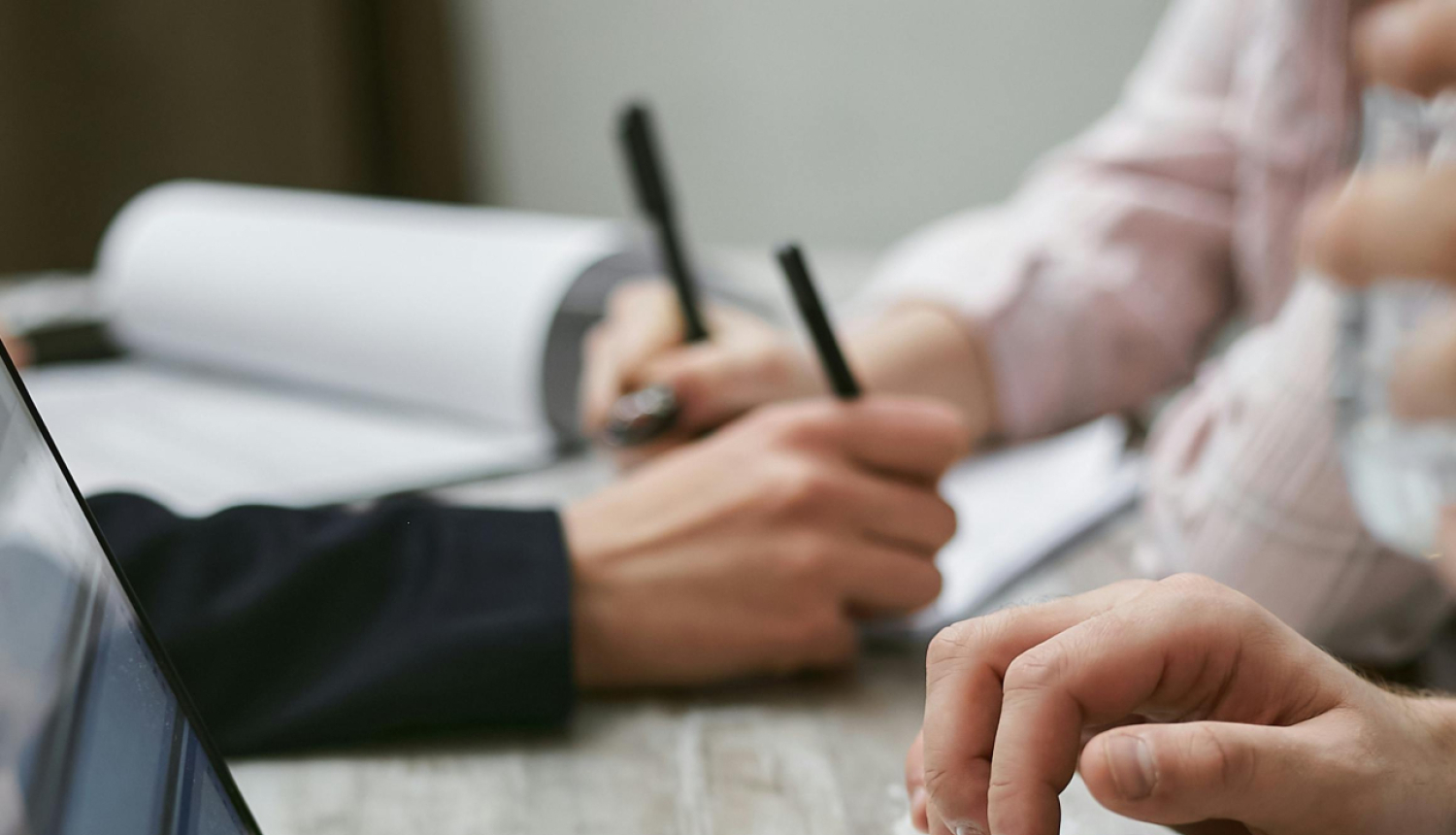 Darba galds ar portatīvo datoru, kā arī redzamas rokas, kas tur pildspalvas