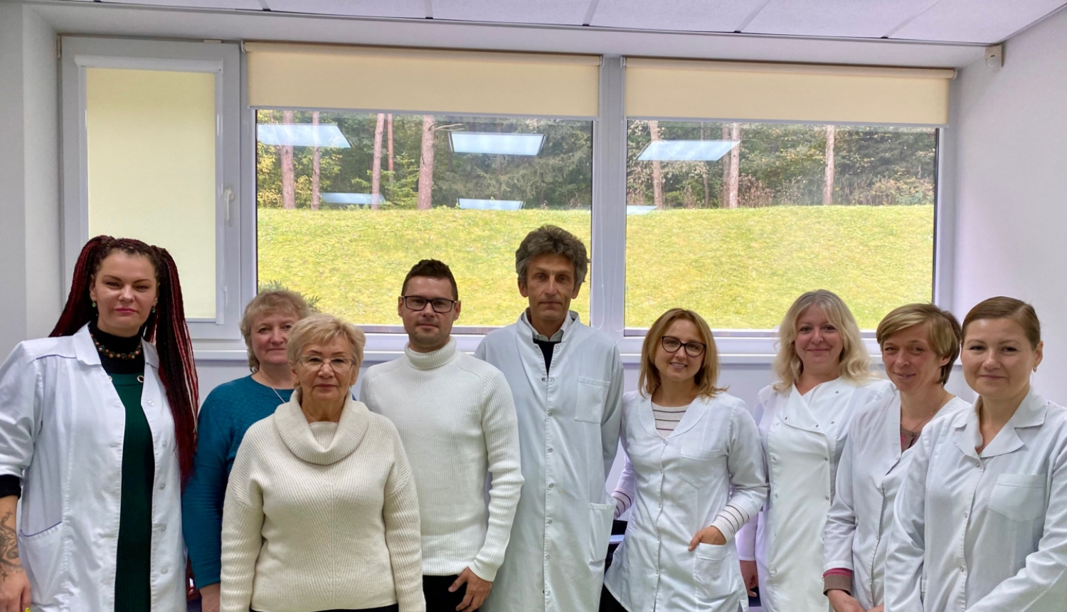 Deviņi cilvēki  baltos halātos stāv ar muguru pret logu