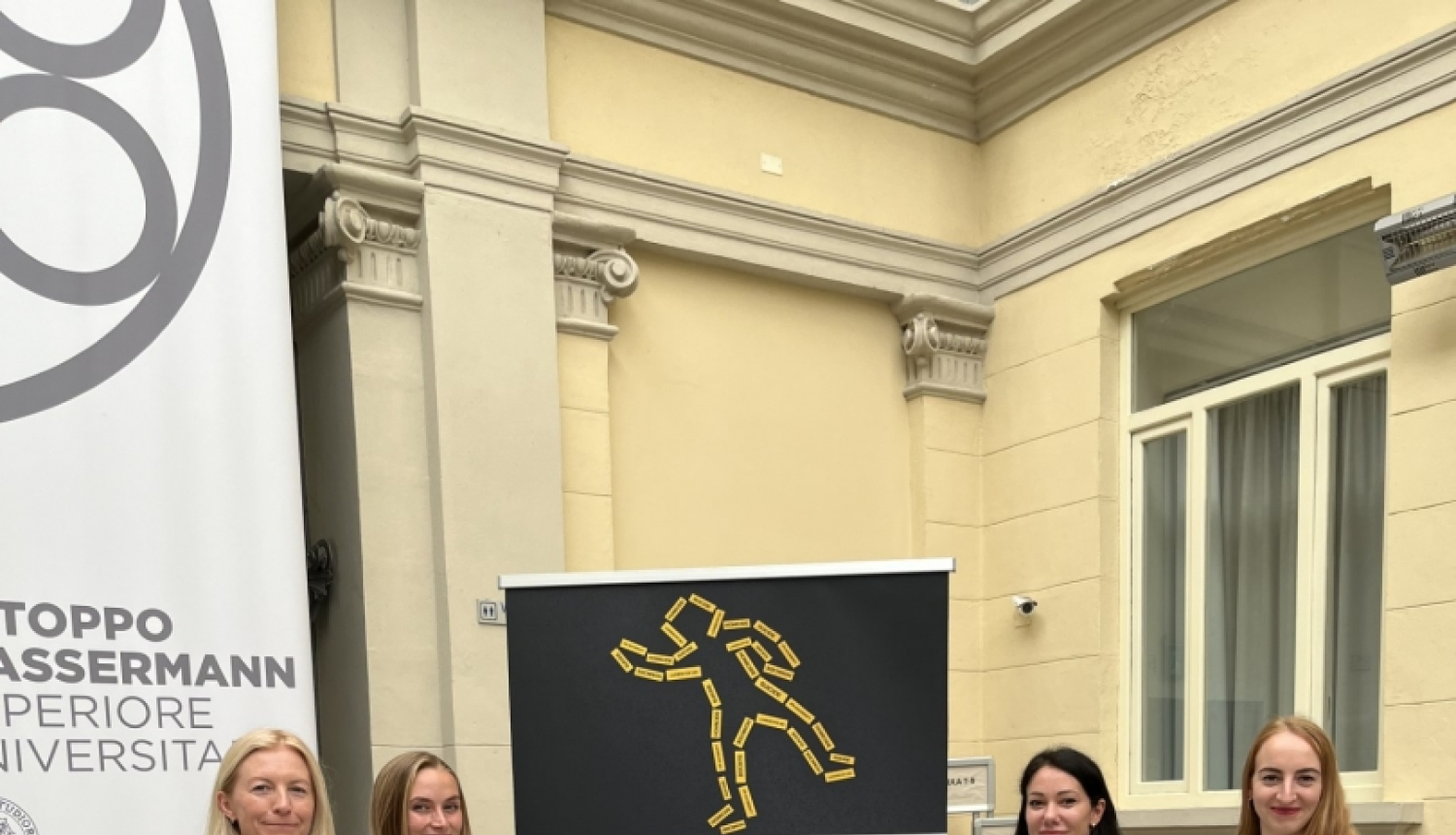 Četras sievietes stāv pie plakāta ar konferences programmu, fonā dzeltena ēka