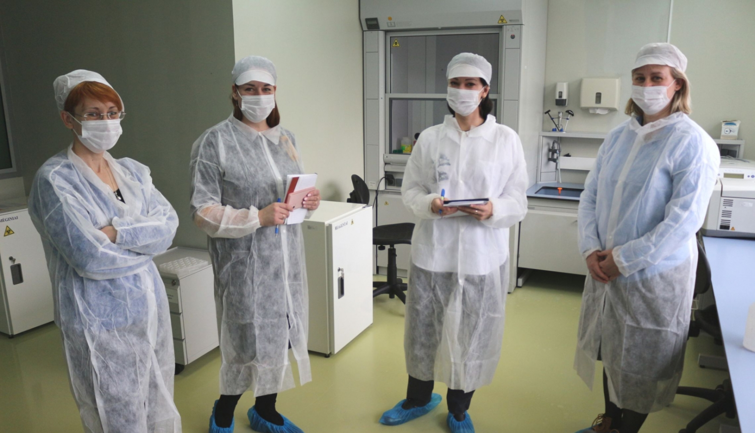 Četras sievietes baltos halātos un ar maskām uz sejas stāv laboratorijas telpās