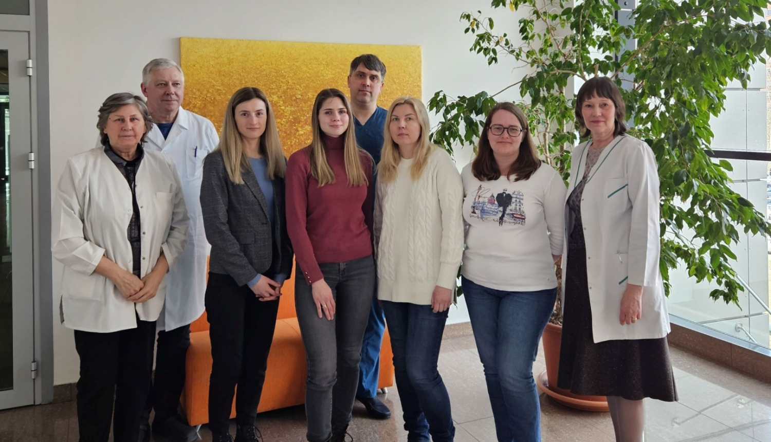 Astoņi cilvēki pozē fotogrāfam, fomā balta siena, liela glezna dzeltenos toņos un liels istabas augs podā