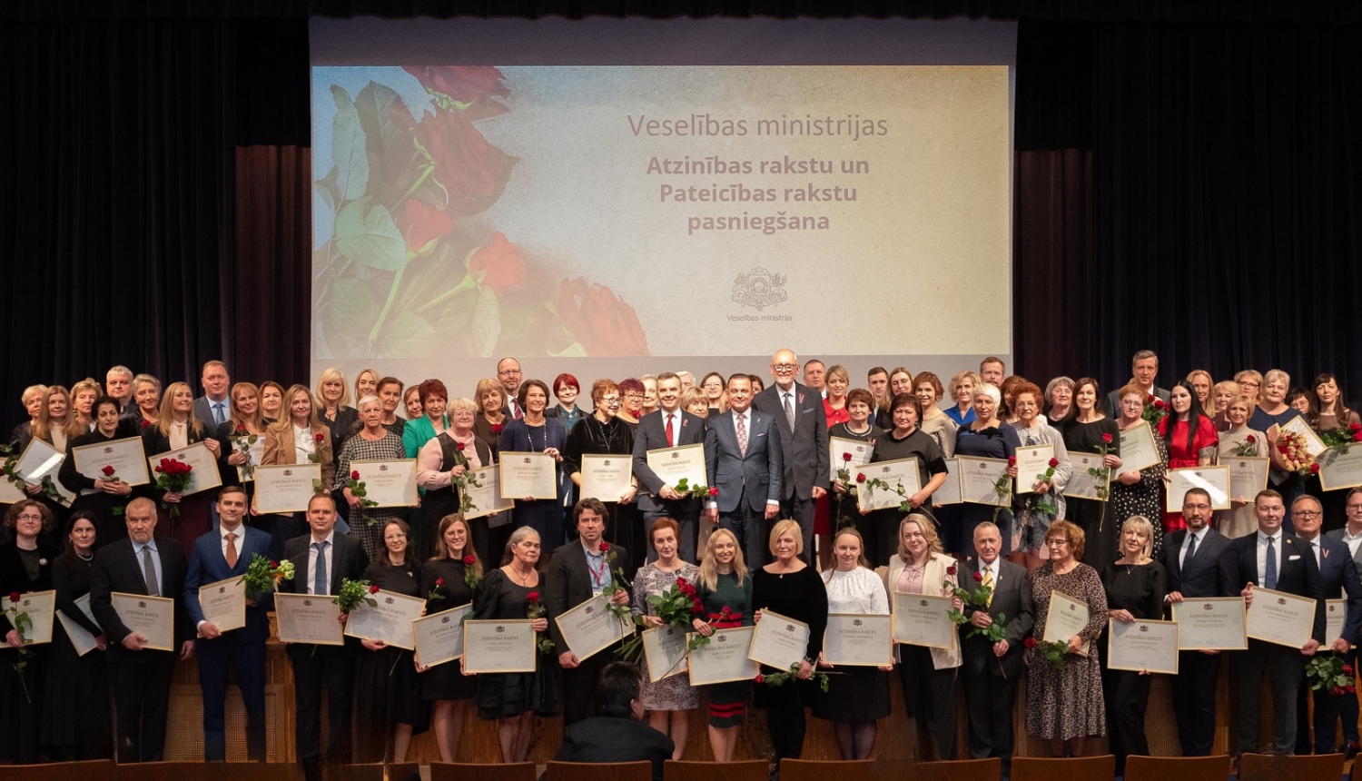 Liels daudzums cilvēku sastājušies vairākās rindās koppbildei, tiem rokās ir ierāmētas atzinības un pateicības raksti