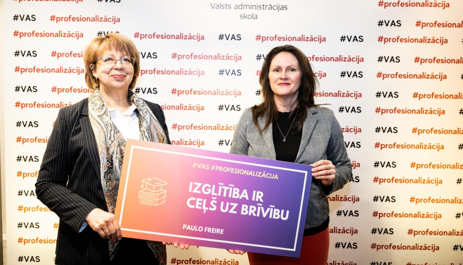 Divas sievietes stāv ar muguru pret sienu, uz kura ģērbonis un uzraksts Valsts administrācijas skola, abas rokā tur plakātu ar uzrakstu - Izglītība ir ceļš uz brīvību